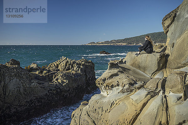 Seitenansicht einer Frau  die auf Felsen am Strand vor klarem Himmel sitzt