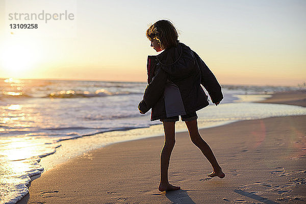 Mädchen in Jacke schaut aufs Meer  während sie bei Sonnenuntergang am Strand steht