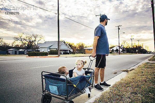 Vater hält Wagen mit Kinderwagen  in dem Kinder auf der Straße sitzen