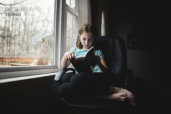 Mädchen lernt in voller Länge  während sie zu Hause auf einem Stuhl am Fenster sitzt
