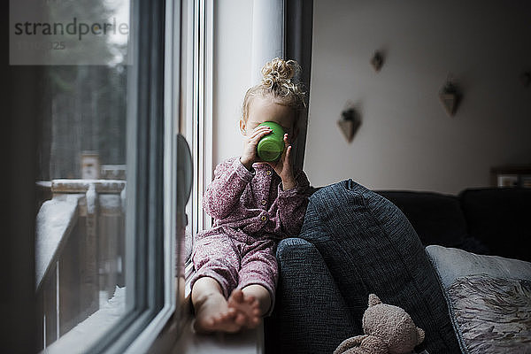 Mädchen  das auf der Fensterbank zu Hause sitzt und etwas trinkt