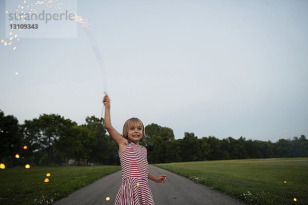 Glückliches Mädchen hält Feuerwerkskörper in der Hand  während sie auf der Straße inmitten eines Grasfeldes im Park steht
