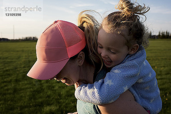 Nahaufnahme einer glücklichen Mutter  die ihre Tochter huckepack nimmt  während sie im Park bei Sonnenuntergang gegen den Himmel rennt