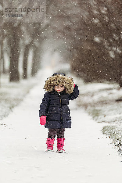 Porträt eines süßen Mädchens mit Kapuzenjacke  das bei Schneefall auf einem verschneiten Fußweg steht