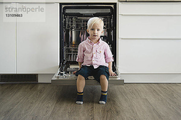 Ganzflächiges Porträt eines süßen Jungen  der zu Hause auf der Tür eines Geschirrspülers sitzt