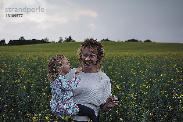 Tochter berührt Blume auf fröhlicher Nase der Mutter auf Feld gegen Himmel