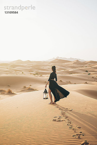 Seitenansicht einer Frau  die eine Laterne hält  während sie bei Sonnenuntergang in der Wüste Sahara vor klarem Himmel steht