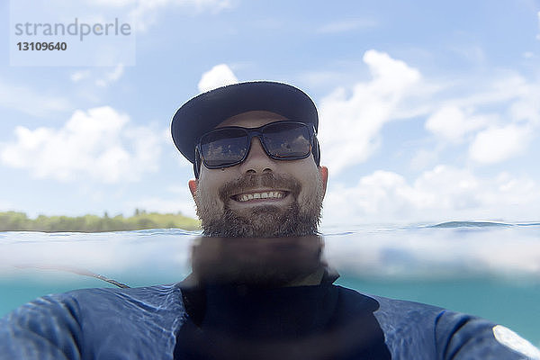 Porträt eines glücklichen Mannes mit Sonnenbrille beim Schwimmen im Meer gegen den Himmel