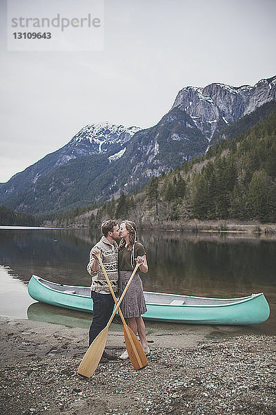 Junges Paar hält Ruder  während es sich am Seeufer im Silver Lake Provincial Park küsst