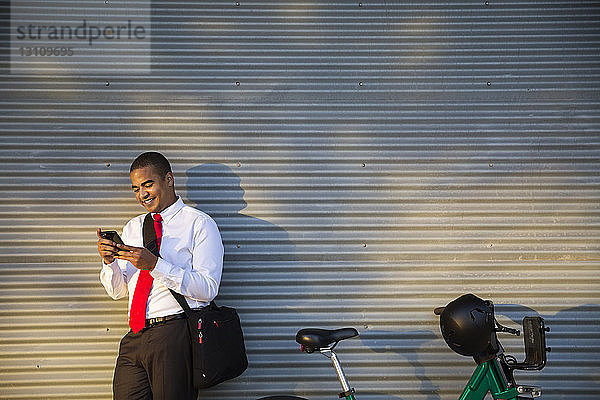 Lächelnder Geschäftsmann  der ein Mobiltelefon benutzt  während er gegen den geschlossenen Verschluss steht