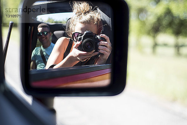 Frau fotografiert sich im Seitenspiegel reflektierend  während sie mit einem Freund im Geländewagen unterwegs ist