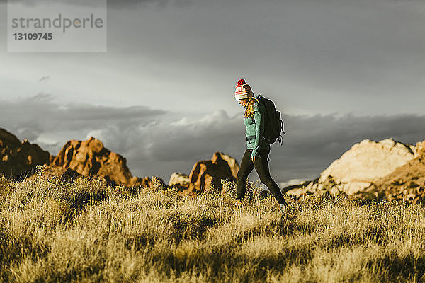 Seitenansicht einer Wanderin mit Rucksack auf einem Grasfeld vor Felsformationen und bewölktem Himmel bei Sonnenuntergang