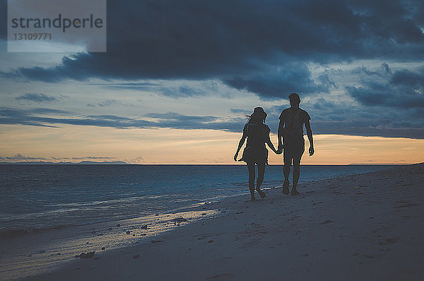 Scherenschnitt-Paar  das sich bei Sonnenuntergang an den Händen hält und am Strand vor bewölktem Himmel spazieren geht