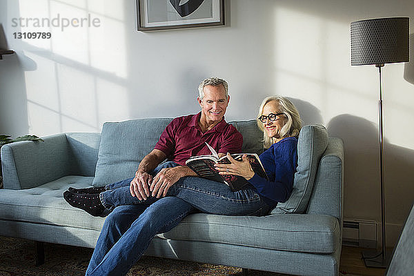 Lächelndes Paar liest Buch  während es sich zu Hause auf dem Sofa im Wohnzimmer ausruht