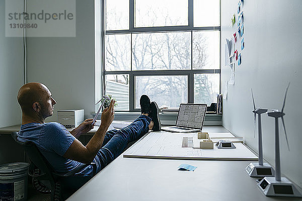 Seitenansicht eines Geschäftsmannes  der das Modell einer Windkraftanlage hält  während er mit den Füßen auf dem Schreibtisch sitzt