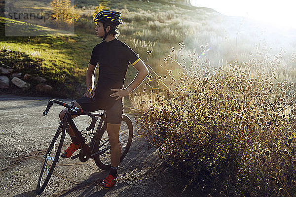 Zuversichtlicher Radfahrer  der bei Sonnenschein mit dem Fahrrad an Pflanzen auf der Straße steht