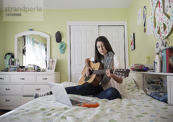 Glückliche Teenagerin spielt Gitarre  während sie zu Hause am Laptop sitzt