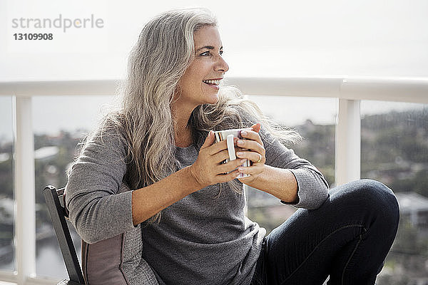 Nachdenkliche reife Frau hält Kaffeetasse  während sie auf dem Balkon sitzt