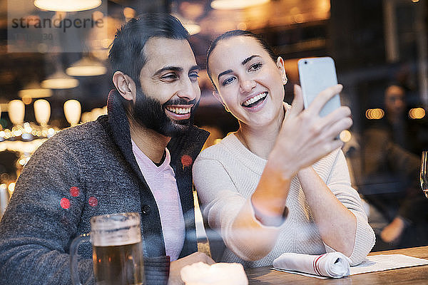 Glückliche Frau mit Freund  der ein Mobiltelefon benutzt  durch Restaurantfenster gesehen