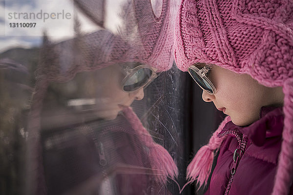 Nahaufnahme eines Mädchens in warmer Kleidung mit Sonnenbrille beim Blick durch das Fenster zu Hause