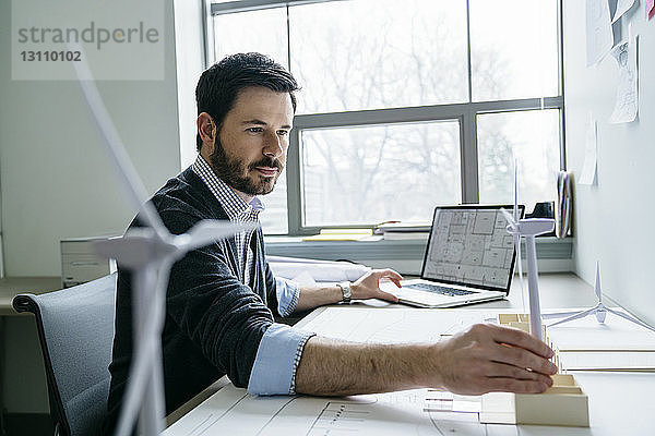 Geschäftsmann benutzt Laptop  während er das Modell einer Windkraftanlage auf dem Schreibtisch arrangiert