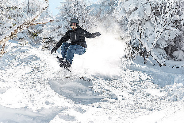 Mann fährt im Winter in voller Länge mit dem Snowboard auf Schnee gegen Bäume