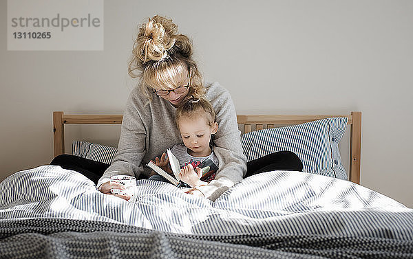 Mutter mit Tochter liest zu Hause am Bett sitzend ein Buch