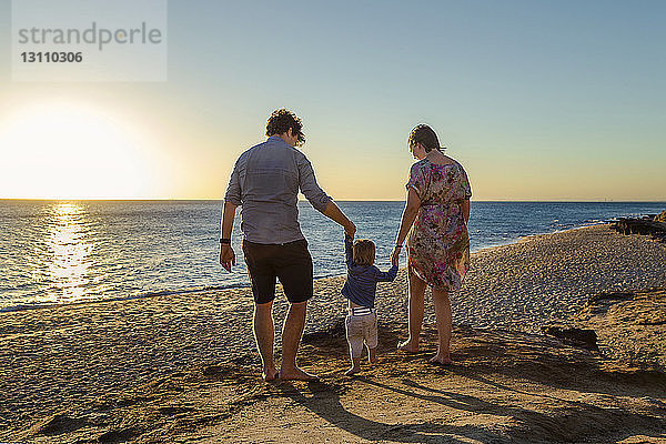 Rückansicht von Eltern  die die Hände des Sohnes halten  während sie bei Sonnenuntergang am Strand spazieren