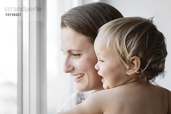 Seitenansicht einer glücklichen Mutter mit Tochter  die zu Hause durchs Fenster schaut