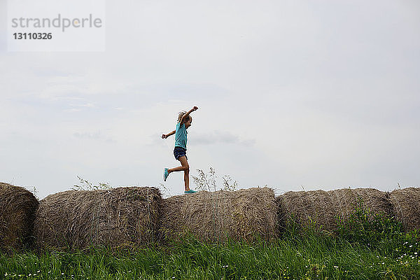 Verspieltes Mädchen rennt auf Heuballen auf Grasfeld gegen den Himmel