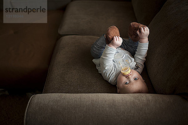Hochwinkelporträt eines kleinen Jungen  der Milch aus der Flasche trinkt  während er zu Hause auf dem Sofa liegt