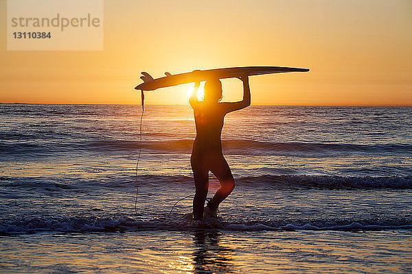 Silhouette einer Surferin  die ein Surfbrett hält und bei Sonnenuntergang am Ufer gegen den Himmel steht