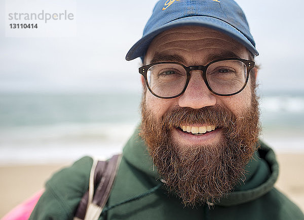 Nahaufnahme eines lächelnden bärtigen Mannes mit Mütze und Brille am Strand