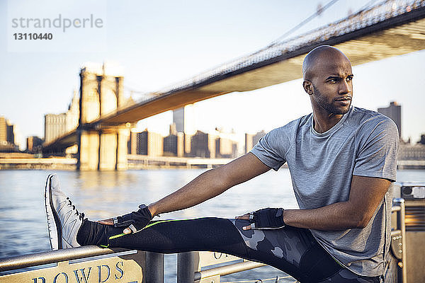 Nachdenklicher männlicher Athlet  der sich auf der Promenade mit der Brooklyn Bridge im Hintergrund das Bein streckt