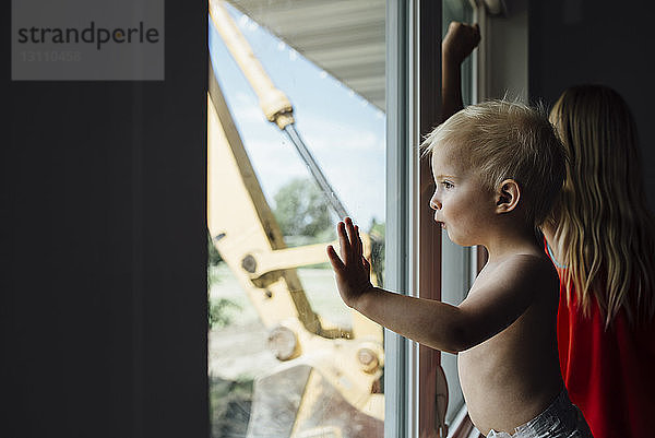 Seitenansicht eines Jungen ohne Hemd  der durch ein Fenster schaut  während er bei seiner Schwester zu Hause steht