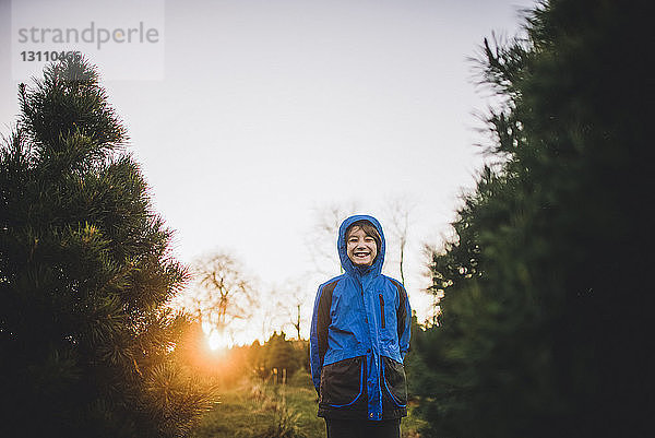 Glücklicher Junge mit Kapuzenjacke  der bei Kiefern auf dem Feld vor klarem Himmel bei Sonnenuntergang steht