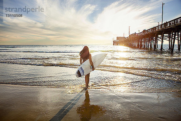 Rückansicht einer Frau  die ein Surfbrett hält und am Ufer steht