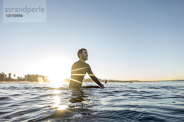 Nachdenklicher Surfer  der bei Sonnenuntergang auf einem Surfbrett im Meer vor klarem Himmel sitzt