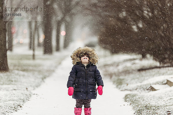 Mädchen mit Kapuzenjacke beim Fangen von Schneeflocken auf der Zunge am verschneiten Fußweg bei Schneefall