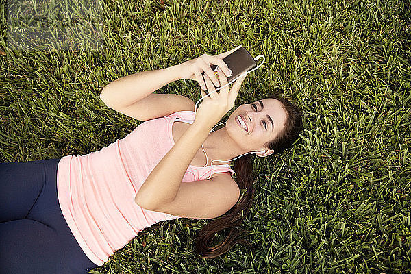 Hochwinkelaufnahme einer glücklichen Frau  die ein Smartphone benutzt  während sie auf einem Grasfeld liegt