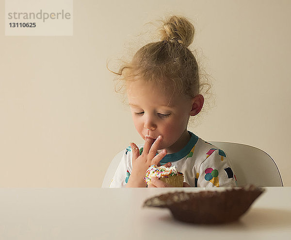 Mädchen leckt Zuckerguss von Törtchen  während sie zu Hause auf einem Stuhl an der Wand sitzt