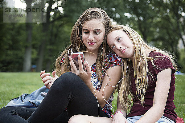 Lächelnde Schwestern hören Musik  während sie sich auf einem Grasfeld im Park entspannen