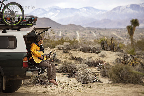 Mann spielt Gitarre  während er auf dem Kofferraum eines Autos sitzt