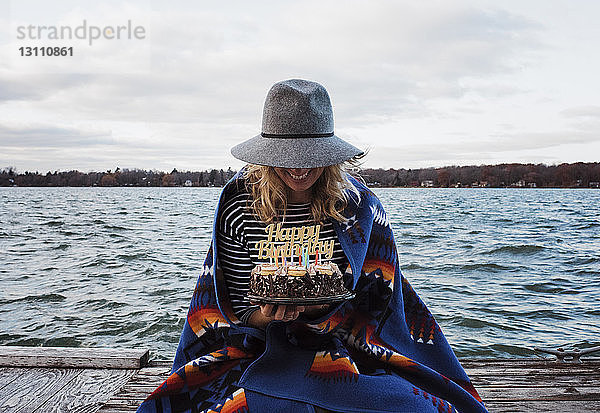 Frau hält Geburtstagskuchen  während sie auf dem Pier am See vor bewölktem Himmel sitzt