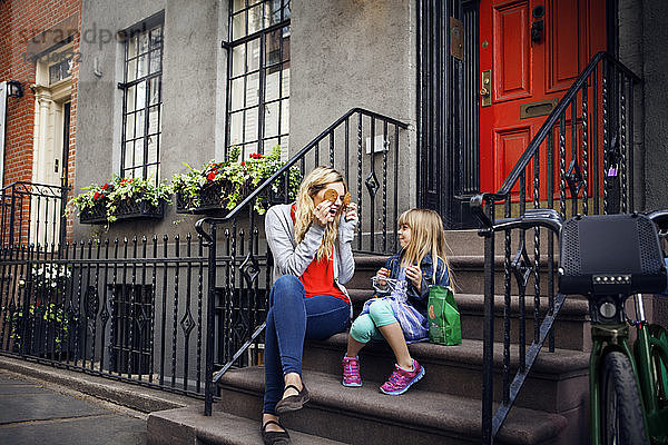 Verspielte Mutter hält Kekse vor die Augen  während sie mit der Tochter auf den Stufen sitzt