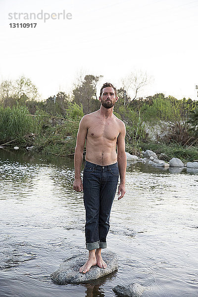 Porträt eines Mannes ohne Hemd  der auf einem Felsen im Fluss vor klarem Himmel steht