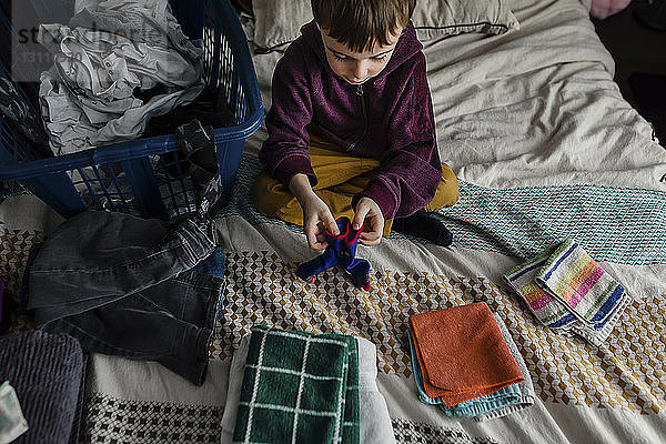 Hochwinkelansicht eines Jungen  der zu Hause auf dem Bett sitzend Kleidung faltet