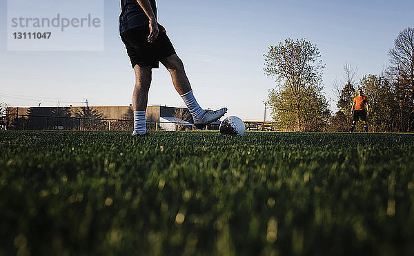 Niedriger Teil eines Mannes  der mit einem Freund auf einem Rasenplatz gegen den klaren Himmel im Park Fussball spielt