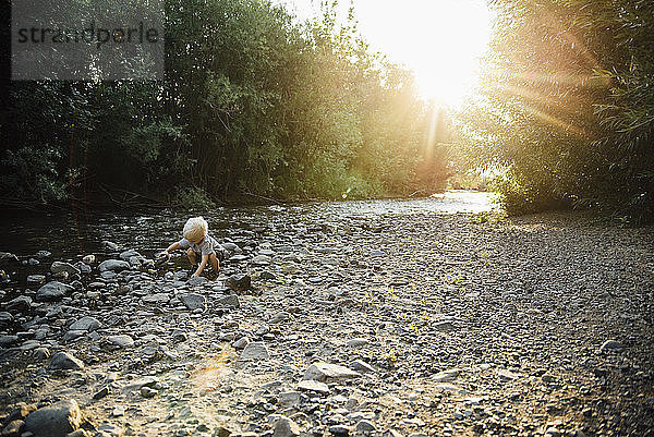 Sorgenfreier Junge spielt mit Kieselsteinen  während er am Fluss kauert