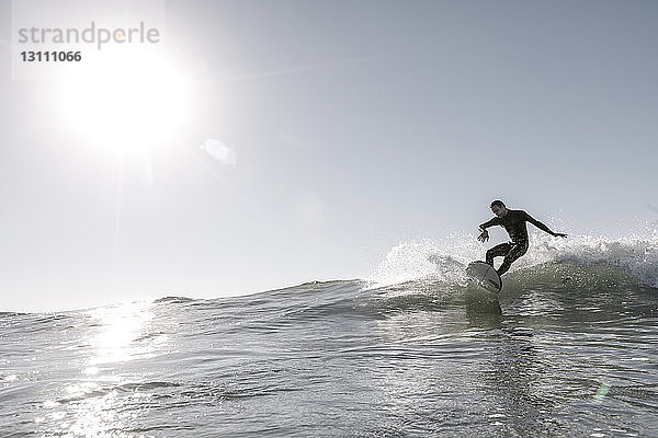 Mittelgroßer erwachsener Mann surft auf riesiger Welle im Meer vor klarem Himmel am sonnigen Tag